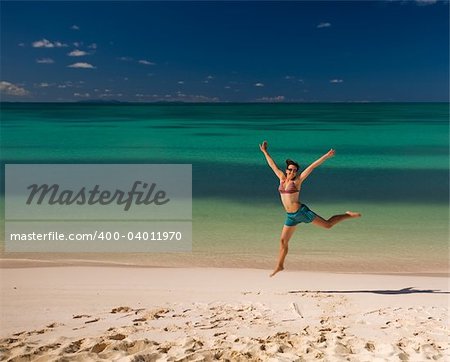 Young woman jumping on white sand beach by turquoise waters on tropical island