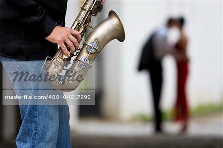 A street musician plays his saxophone while a romantic couple can be seen out of focus in the background