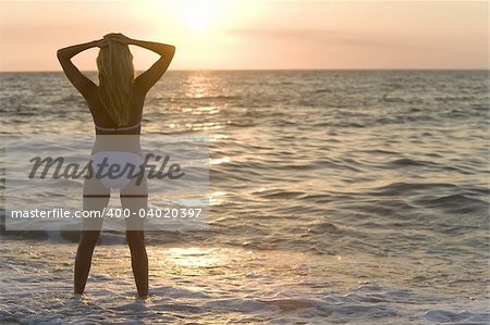 A beautiful bikini clad blond stands in the surf at sunset