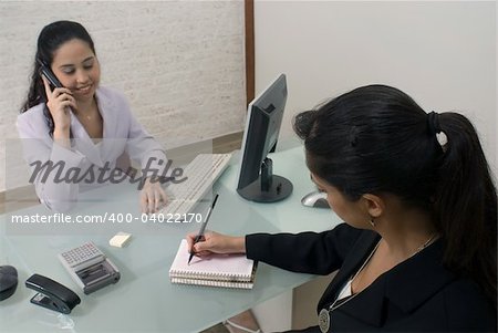 Two latin american businesswomen in a meeting. One is on the phone, the other is taking notes