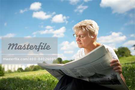The woman in old age sits on a grass in park on a background of the blue sky and reads the newspaper