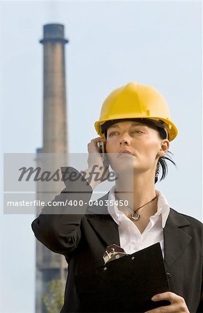 A beautiful dark haired woman wearing a hard hat and talking on her mobile phone while standing in front of a factory and its chimneys