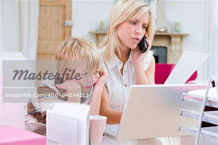 Woman using telephone in home office with laptop while young boy