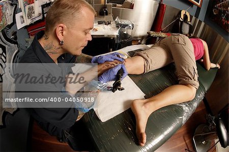 A tattoo artist applying his craft onto the lower leg of a pain-inflicted female.