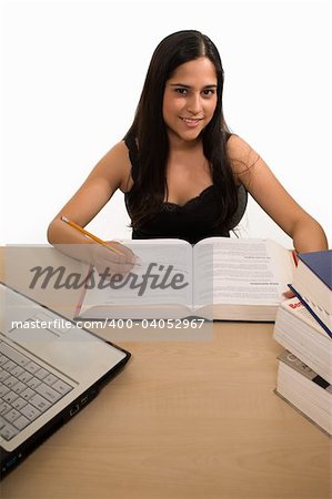 Young Hispanic woman sitting in front of desk with a pile of thick textbooks while reading one