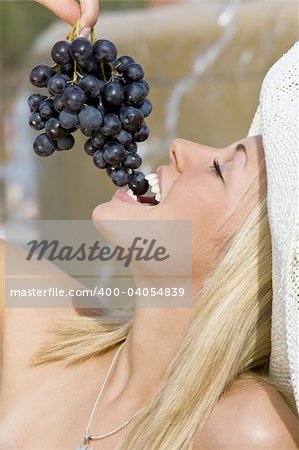 A beautiful young blond woman sitting in front of a fountain eating a bunch of black grapes