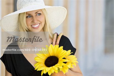 A beautiful young blond woman wearing a sun hat and smiling while carrying a shopping bag of sunflowers