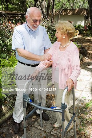 Senior couple outdoors.  She's in a walker and he's helping her.