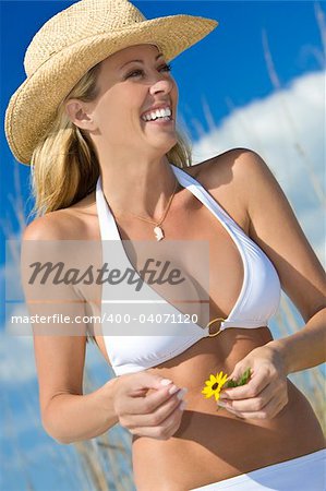 A beautiful blond haired blue eyed model wearing a white bikini laughs while holding a flower amid tall grass