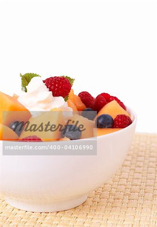 Bowl of summer fruit salad with raspberries, melon, blueberries, cream and mint leaves in a white bowl isolated