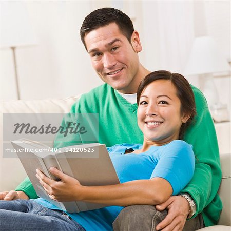 An attractive young couple sitting on a couch together with a book.  The female is sitting in the male's lap.  They are smiling directly at the camera. Square framed shot.