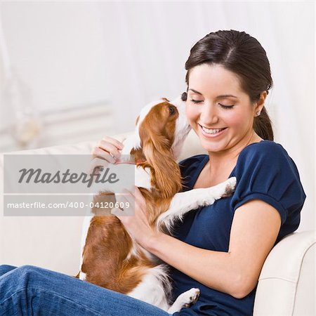 An attractive young woman sitting on a couch and being kissed by a dog that she is holding.  She is smiling and her eyes are closed. Square framed photo.