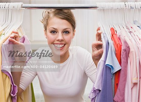 A young, attractive woman is standing in a closet and looking through clothes.  She is smiling at the camera.  Horizontally framed shot.