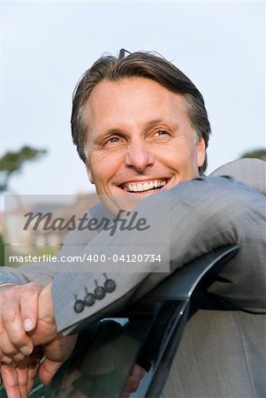 A colour portrait of a handsome smiling forties businessman leaning on the door of his car and looking to the side.