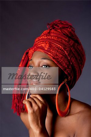 Belle femme afro-américaine vêtu d'un traditionnel tribal rouge orange foulard sur la tête et big orange créoles, appuyant son menton avec une main, isolé.
