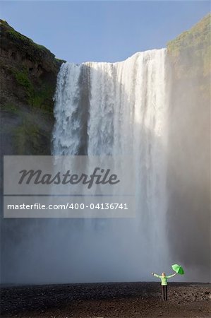 A woman dressed in a green jacket carrying a green ubmrella standing arms outstretched at the base of a waterfall. Shot on location at Skogafoss in Iceland.