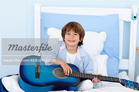 Happy little boy playing guitar sitting in bed