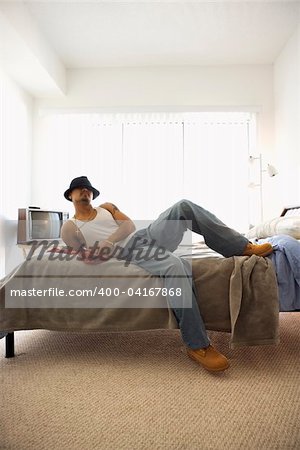 A young man lying on a bed. He is dressed in casual clothing and wearing a hat. Vertical shot.