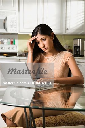 A young woman sits at the kitchen table using a laptop. She has a worried expression on her face. Vertical shot.