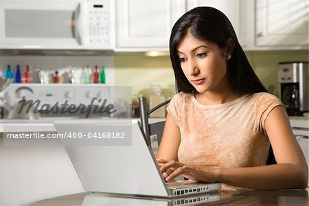 A young woman sits at the kitchen table while using a laptop. Horizontal shot.