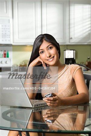 Portrait of a young woman sitting at the kitchen table with a laptop and a cell phone. Vertical shot.