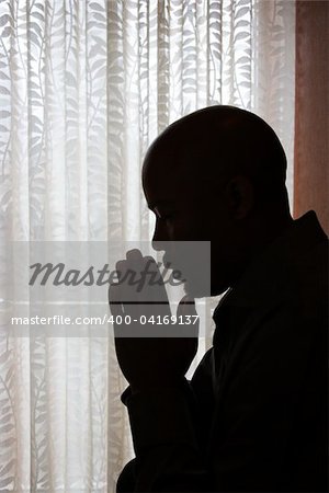 Profile of African-American man silhouetted by a window with his head bowed and his hands in prayer. Vertical shot.