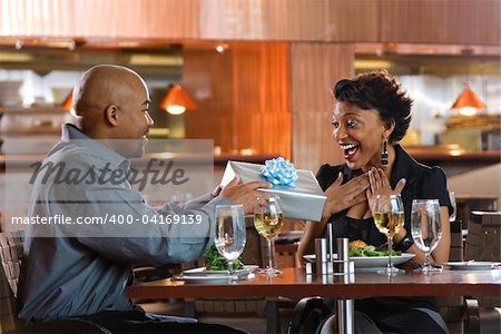 African-American man giving a surprised woman a gift-wrapped present at a restaurant. Horizontal shot.