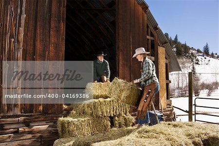 Attractive young man and woman lifting hay bales on a farm in front of a barn. Horizontal shot.