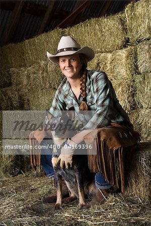 Attractive young woman wearing a white cowboy hat. She is sitting on hay and smiling while holding an Australian Shepherd. Vertical shot.