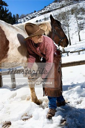 Man wearing a cowboy hat cleans out a horse hoof in the snow. Vertical shot.