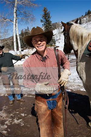 Man in a cowboy hat smiles next to a horse. Another man is standing in the background. Vertical shot.