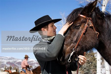 Attractive young man wearing a cowboy hat. He is petting a horse with another rider in the background. Horizontal shot.