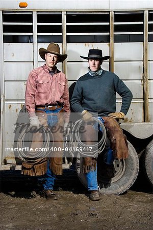 Two men wearing cowboy hats and leaning on the side of a livestock trailer. They are holding lariats. Vertical shot.