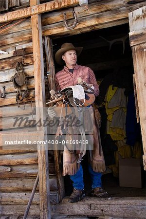 Man wearing a cowboy hat, leaning in the doorway of log cabin and holding a saddle. Vertical shot.