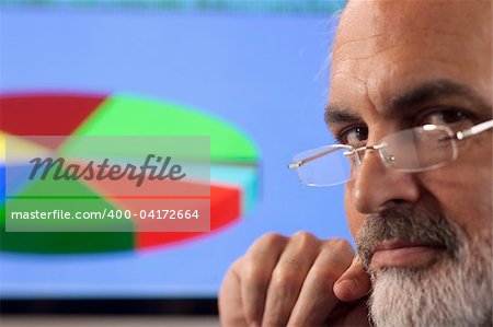 Close-up portrait of a businessman looking pensively at the camera. A computer monitor displaying a multi-colored pie chart can be seen in the background. Horizontal shot.