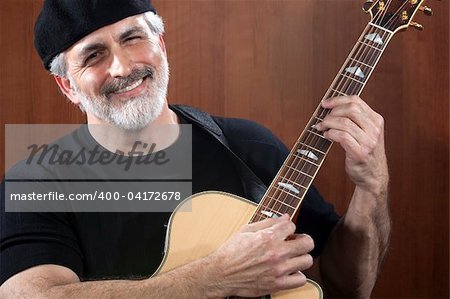 Portrait of a middle-aged man wearing a black beret and t-shirt and playing an acoustic guitar. He is smiling at the camera. Horizontal shot.