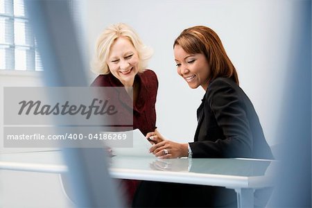 Businesswomen sitting at office desk discussing paperwork smiling.