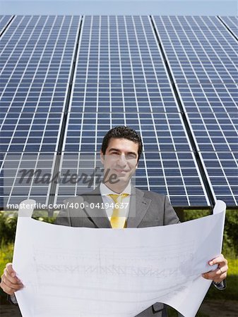 Portrait of mid adult italian male engineer reading blueprints in solar power station and smiling. Vertical shape, front view. Copy space