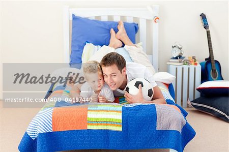 Attractive father and his son playing with a soccer ball lying on bed