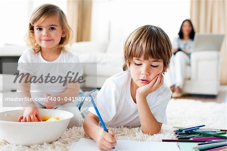 Serious little boy drawing and his sister eating chips lying on the floor