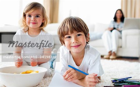 Smiling siblings eating chips and drawing lying on the floor