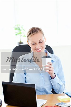 Smiling businesswoman holding a coffee while using a laptop at work