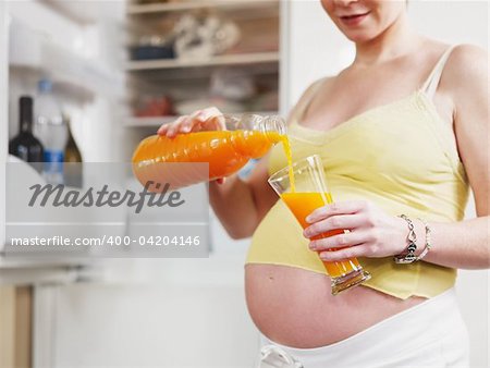cropped view of italian 6 months pregnant woman standing near refrigerator and drinking orange juice. Horizontal shape