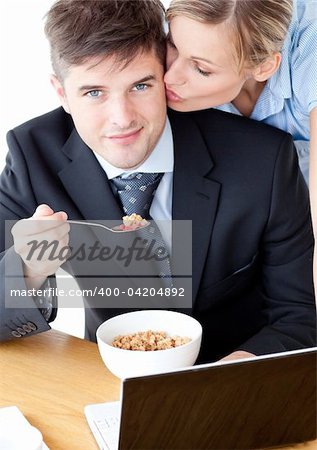 Smiling businessman eating breakfast using laptop while wife kissing his cheek