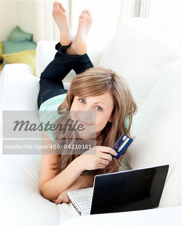 Smiling woman lying ona sofa holding a card and laptop in the living room