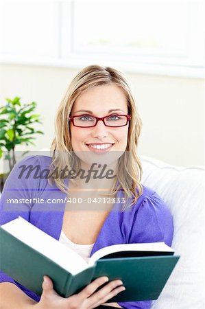 Smiling young woman wearing red glasses reading a book on a sofa at home