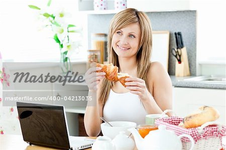 Smiling woman having breakfast in front of the laptop in the kitchen at home