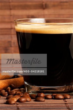 espresso in a short glass with coffee beans and cinnamon sticks from side on wooden background