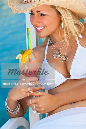 Stunningly beautiful young blond woman in straw cowboy hat and white bikini enjoying a cocktail by a turquoise blue sea