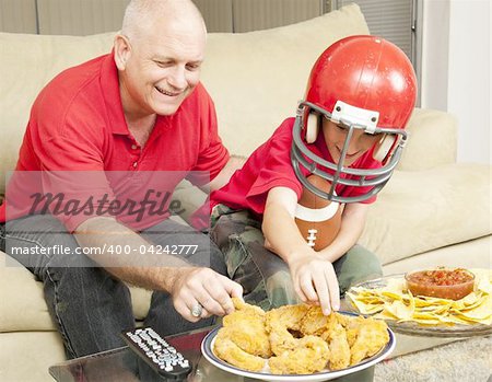 Father and son watching football together and eating snacks.